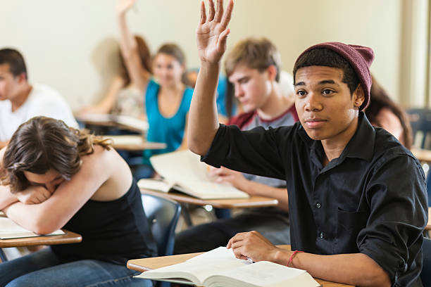 A high school student raises his hand while others work on schoolwork in a classroom of diverse teen students.